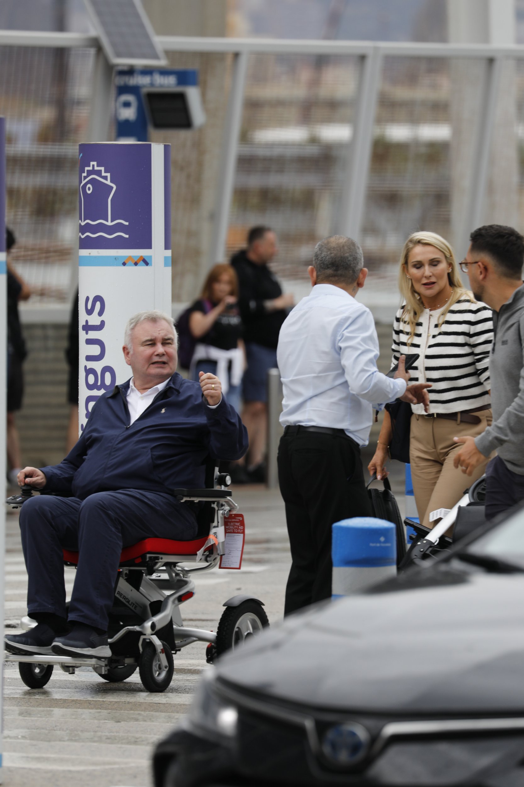 Eamonn Holmes wearing a navy blue jacket and trousers and using a wheelchair at Barcelona airport
