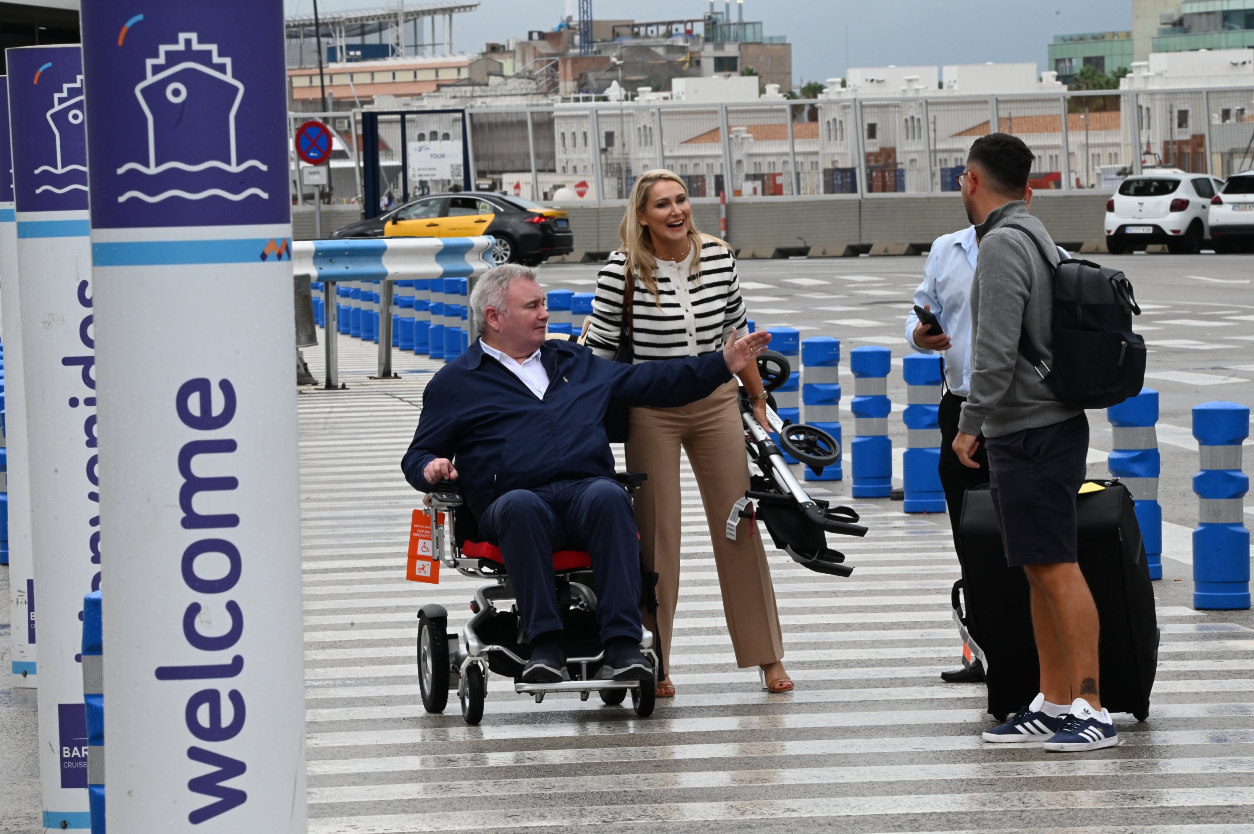 Eamonn Holmes sticking his arm out while in a wheelchair at Barcelona Airpot next to Katie Alexander